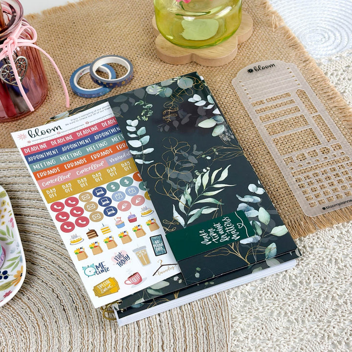A styled flat-lay of a Bloom planner sticker set and matching planner accessories on a burlap mat. The main product is a dark green floral sticker sheet with white botanical illustrations, placed next to a clear plastic planner divider and a colorful planner sticker sheet showing various labels and icons.