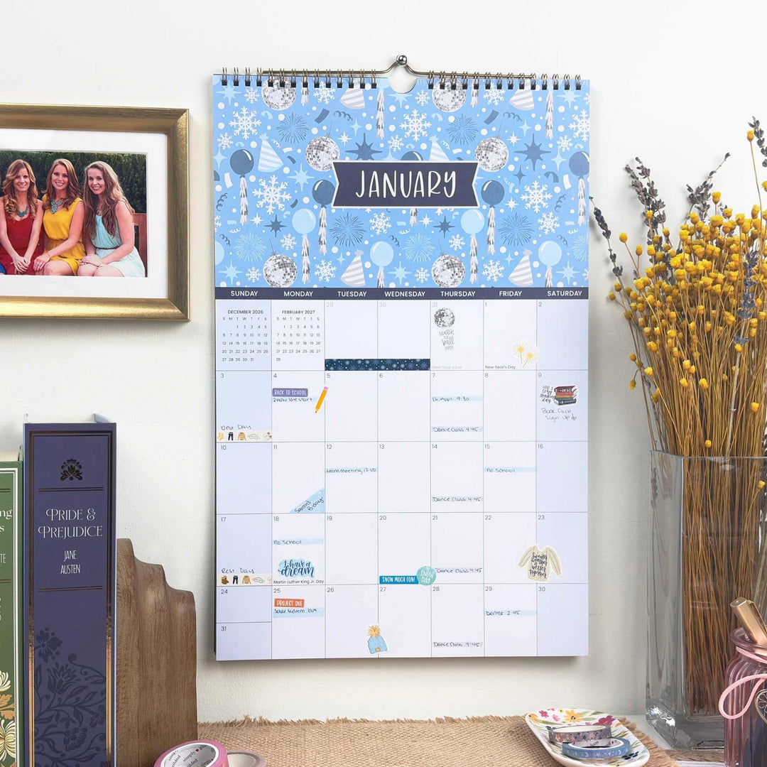 Wall calendar with a winter-themed design on a wall next to a vase with yellow flowers and a framed photo.
