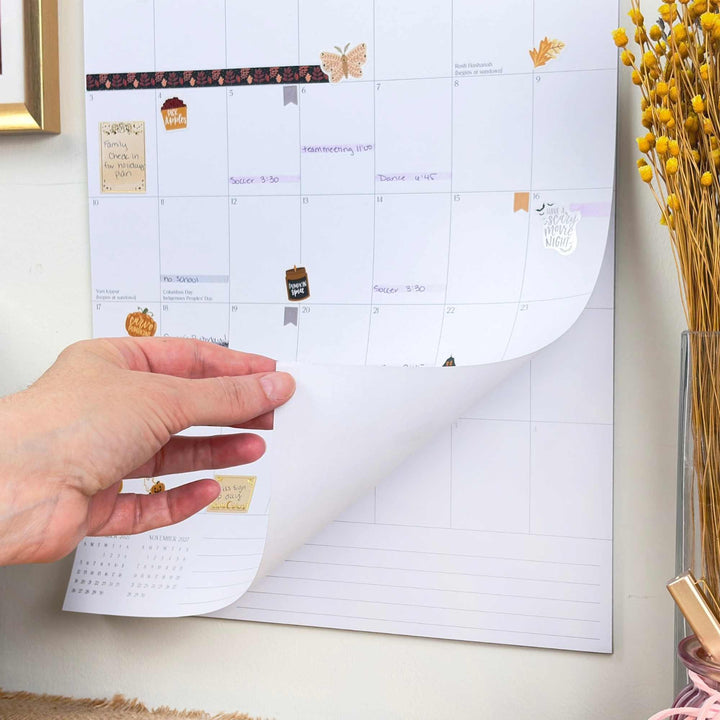 Hand rolling out a wall calendar with decorative stickers on a light-colored wall.