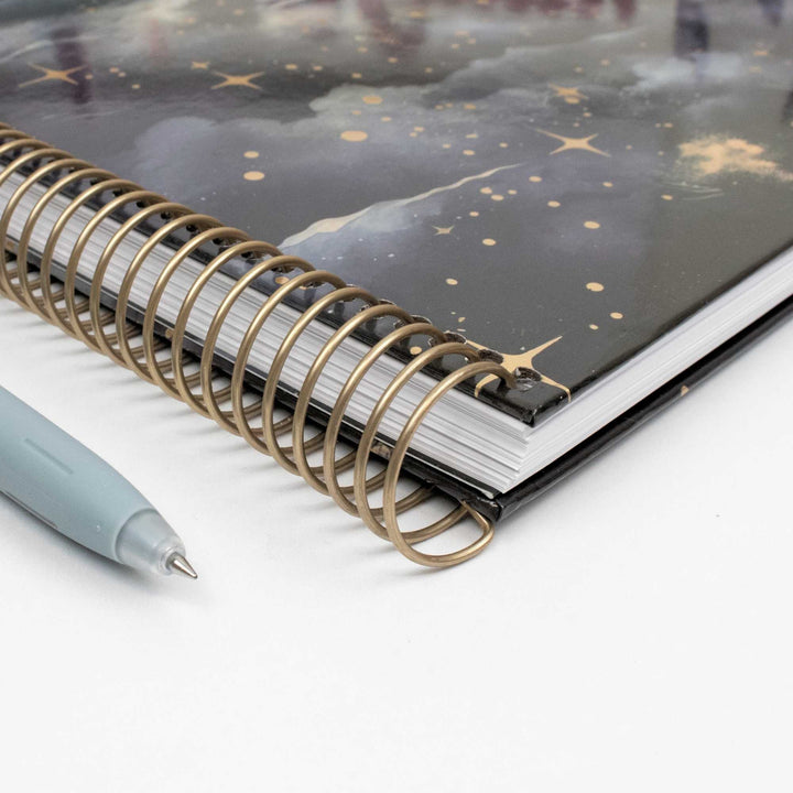A close-up of a spiral-bound notebook with a black cover decorated with gold stars and gold glittery accents, shown on a white surface next to a light blue pen.