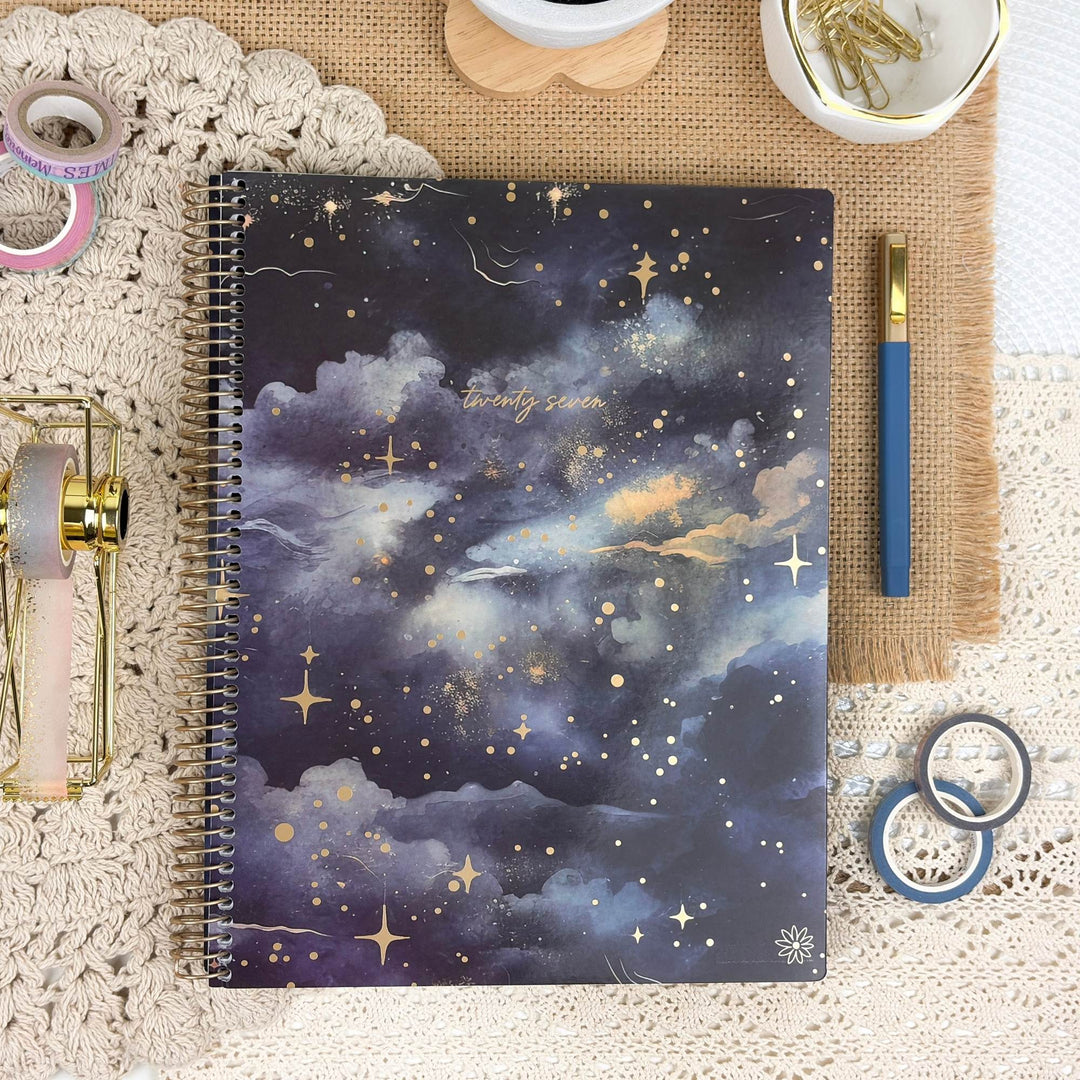 A spiral-bound notebook with a dark blue and purple galaxy cloud cover, styled in a flat-lay arrangement with a blue pen, washi tapes, and stationery accessories on a lace and burlap background.