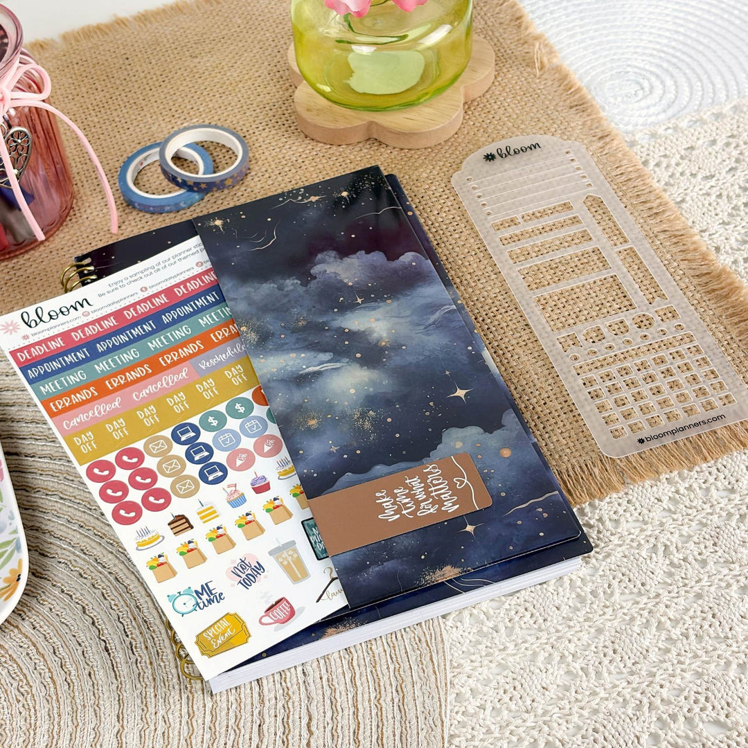 A planner sticker sheet and matching planner accessories are arranged on a woven beige mat. The main product is a dark blue night-sky themed planner cover with stars and clouds, accompanied by a sheet of colorful planner stickers and a clear plastic date-grid template.