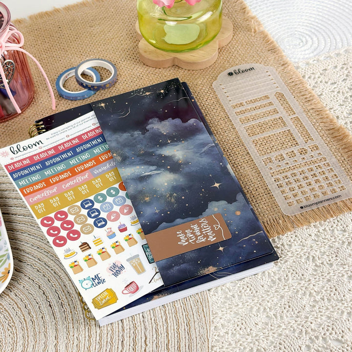 A planner sticker sheet and matching planner accessories are arranged on a woven beige mat. The main product is a dark blue night-sky themed planner cover with stars and clouds, accompanied by a sheet of colorful planner stickers and a clear plastic date-grid template.