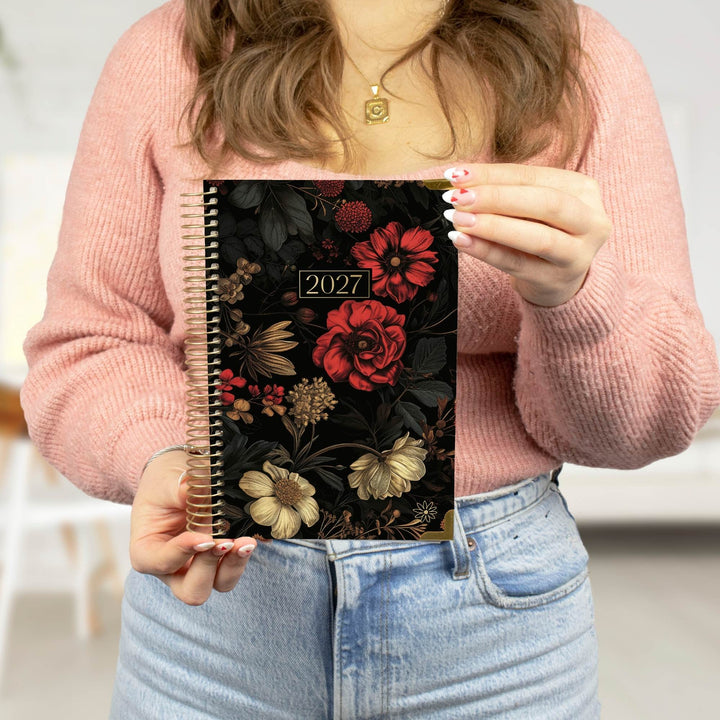 A person in a pink sweater holds a 2027 spiral-bound planner with a black cover decorated with red and gold floral illustrations.