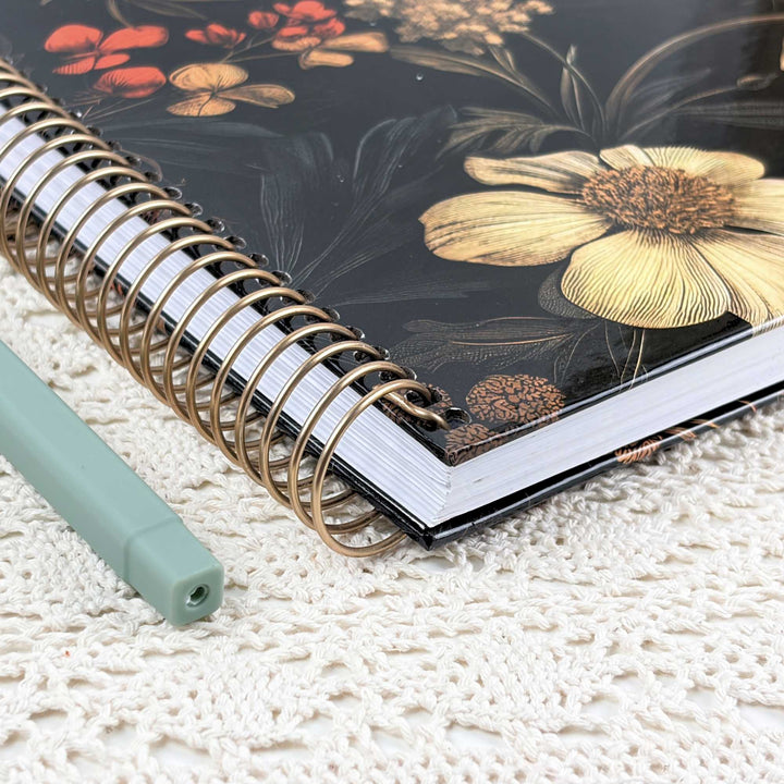 A spiral-bound notebook with a black cover decorated with a large yellow flower and smaller red and gold floral motifs, placed on a white textured knit surface next to a mint green pen.