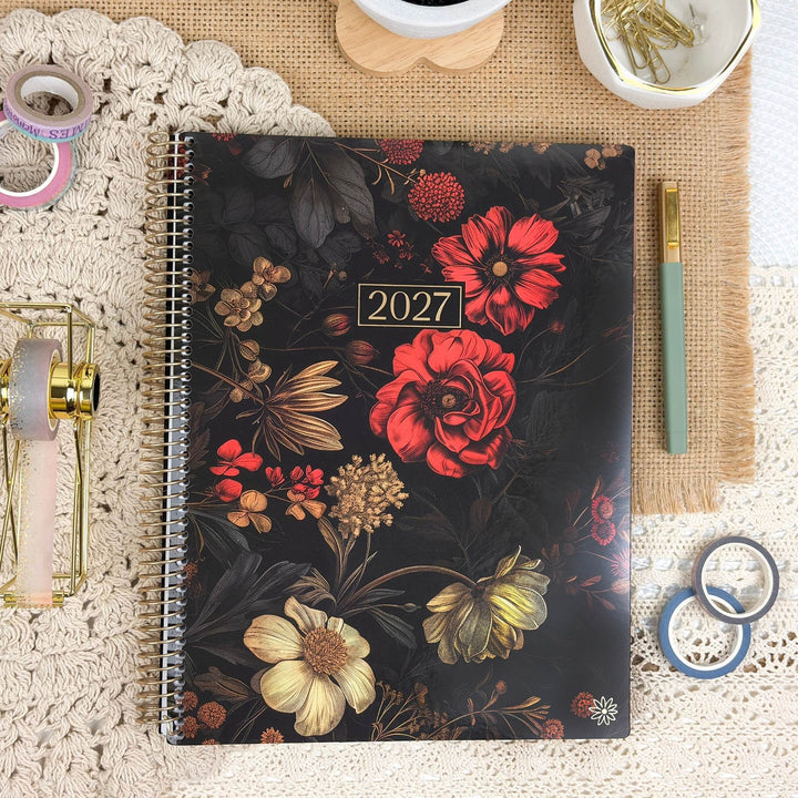 A 2027 spiral-bound planner with a dark floral cover lies on a lace doily surrounded by stationery props including washi tapes, a gold pen, rings, and a small dish of paper clips.