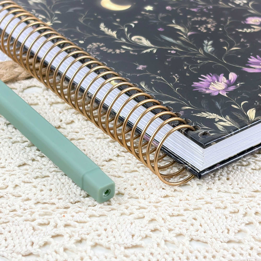 A spiral-bound notebook with a black floral cover and gold wire spirals, placed on a cream lace surface next to a mint green pen.
