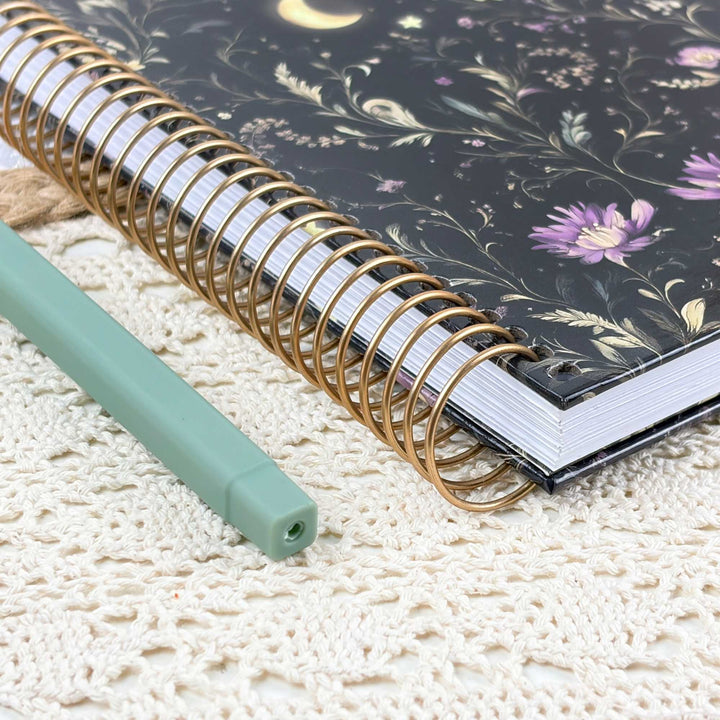 A spiral-bound notebook with a black floral cover and gold wire spirals, placed on a cream lace surface next to a mint green pen.