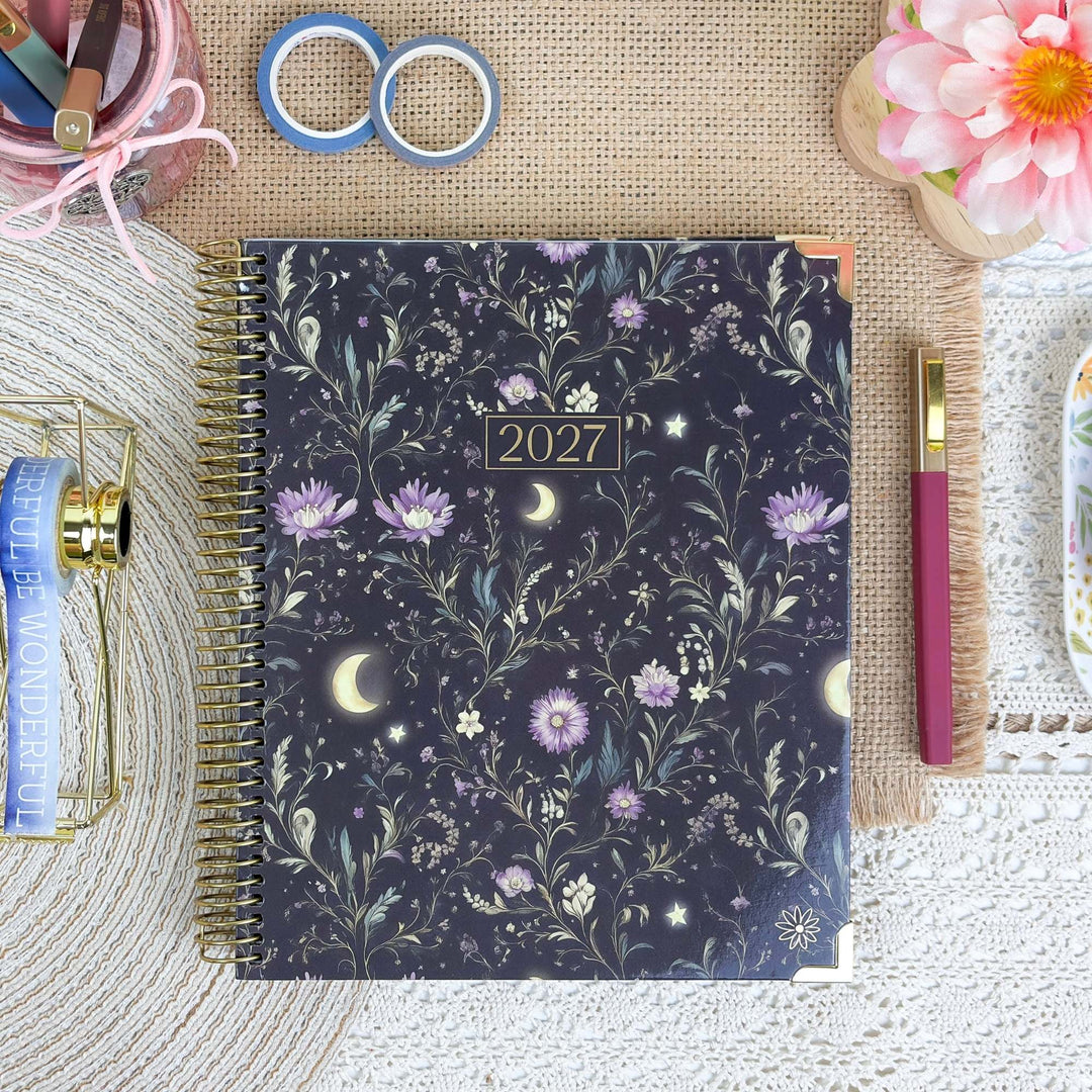 A spiral-bound 2027 planner with a dark navy floral and celestial pattern, styled in a flat-lay arrangement with a pink pen, washi tapes, a small basket, and decorative props on a burlap and lace surface.