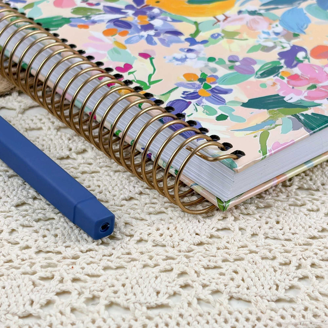 A spiral-bound notebook with a colorful floral cover lies on a cream lace doily next to a blue pen.