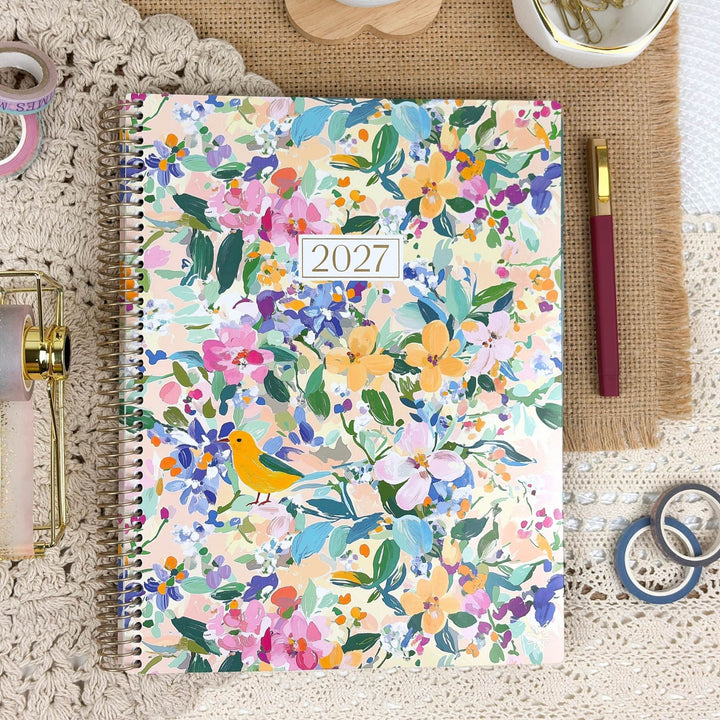 A 2027 spiral-bound planner with a colorful floral cover design, styled in a flat-lay arrangement on a lace doily with stationery props.
