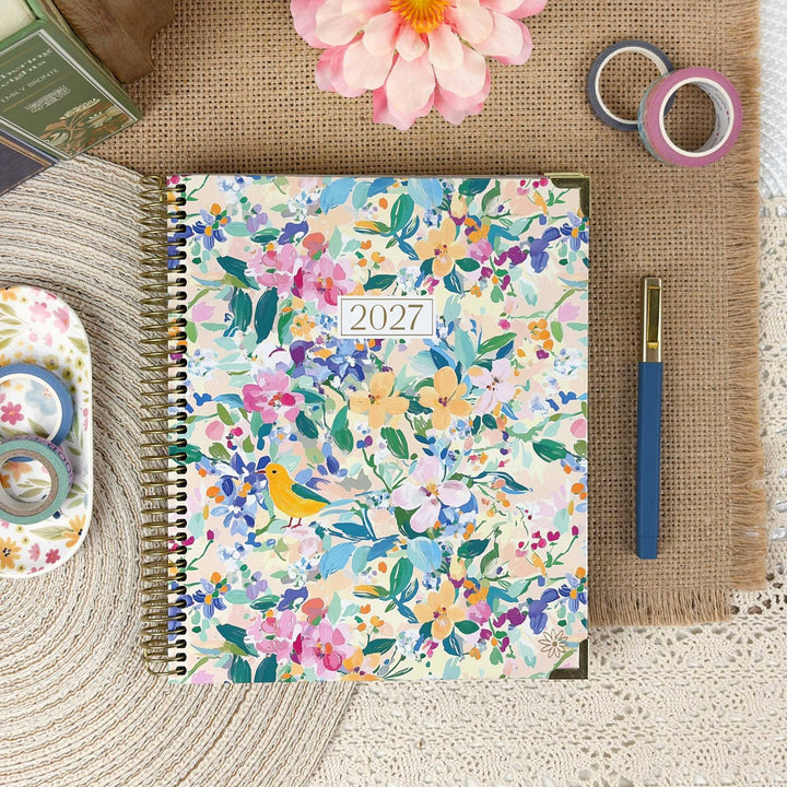 A spiral-bound 2027 floral planner lies on a burlap mat, surrounded by stationery props including a blue pen, washi tapes, a pink flower, and a small decorative dish.