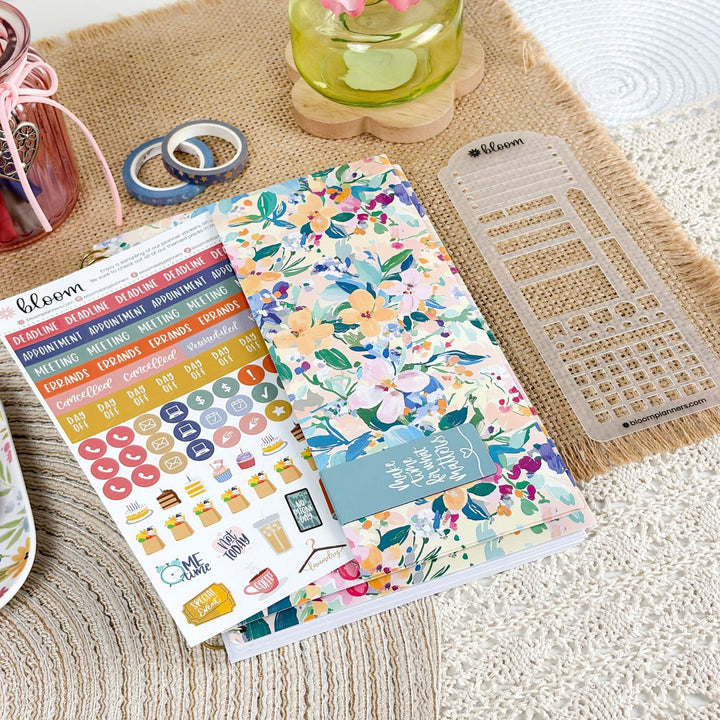 A floral planner sticker sheet and matching planner accessories arranged on a woven beige mat, with a small glass jar, washi tape, and a clear planner grid visible in the scene.