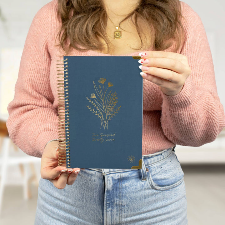 A person in a pink knit sweater and light blue jeans holds a navy blue spiral-bound planner or notebook with gold floral line art and gold text on the cover.