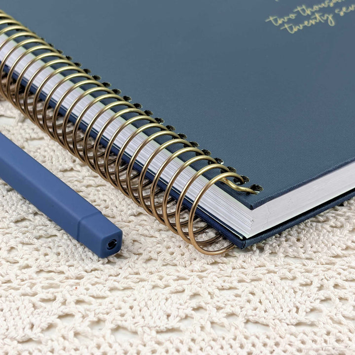 A navy blue spiral-bound notebook with a gold wire coil binding, placed diagonally on a cream-colored textured lace doily next to a matching blue pen.