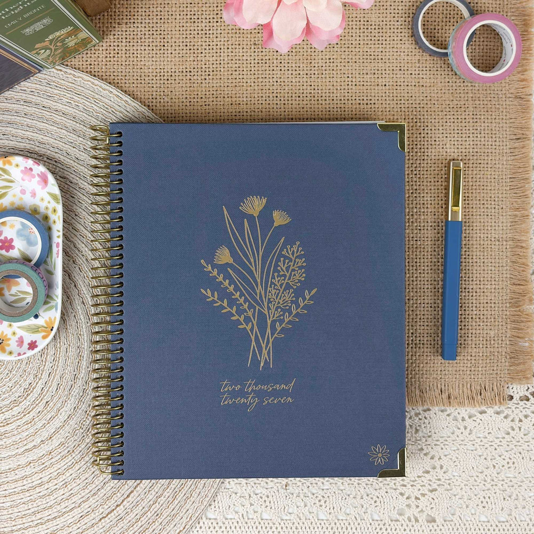A navy blue spiral-bound planner or notebook with gold floral line art and the text 'two thousand twenty seven' on the cover, styled on a burlap mat with a blue pen, washi tapes, and decorative paper around it.