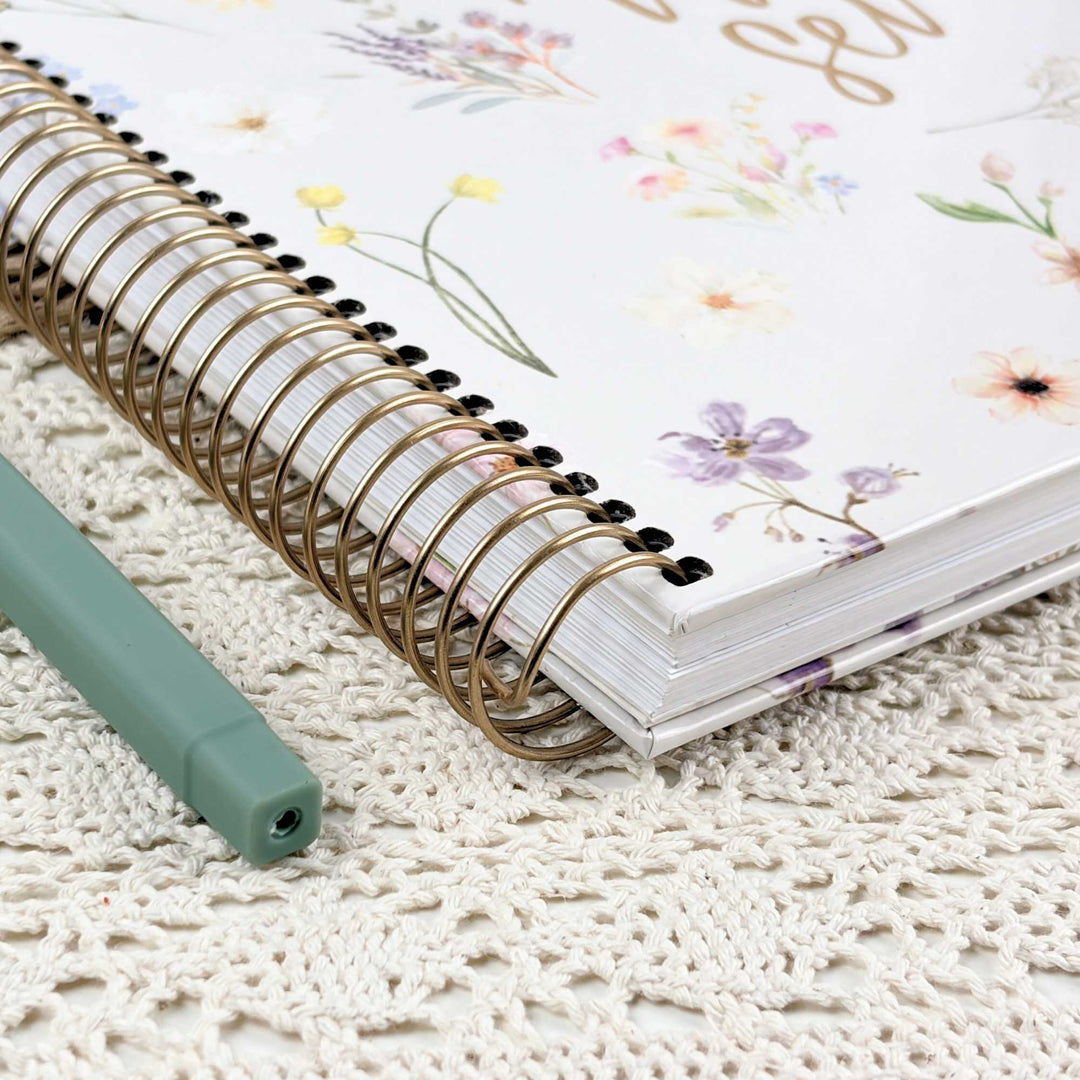 A floral-bound spiral notebook with a gold-tone wire binding and a white cover decorated with pastel flowers, shown on a cream lace surface next to a mint green pen.