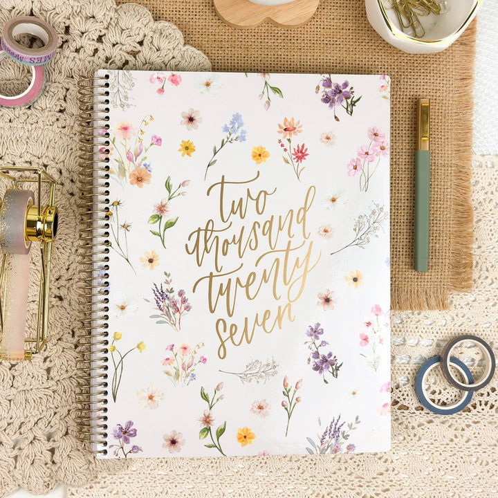 A spiral-bound planner or notebook with a white cover decorated with pastel watercolor flowers and gold script reading 'Two thousand twenty seven,' styled on a crocheted surface with pens, washi tape, and small decorative items around it.