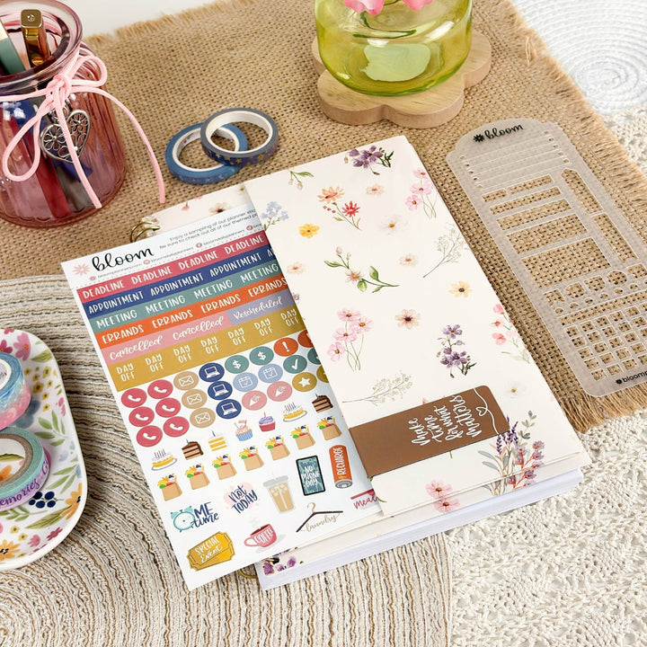 A styled flat-lay of planner sticker sheets and a floral planner notebook on a burlap mat, surrounded by stationery props including washi tape, a pen cup, and a clear plastic divider.