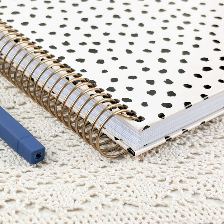 A spiral-bound notebook with a white cover patterned with black spots, resting on a cream-colored crocheted textile next to a blue pen.