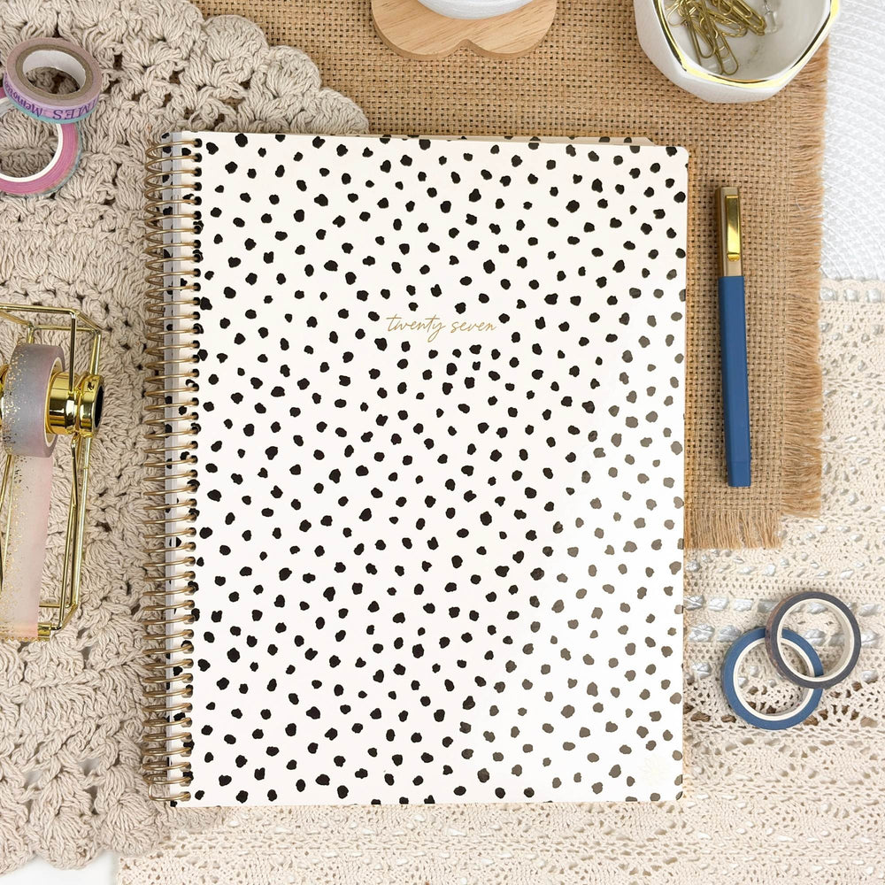 A spiral-bound planner or notebook with a white cover covered in small black polka dots, shown in a styled flat-lay with a blue pen, washi tapes, and other stationery around it. - hover