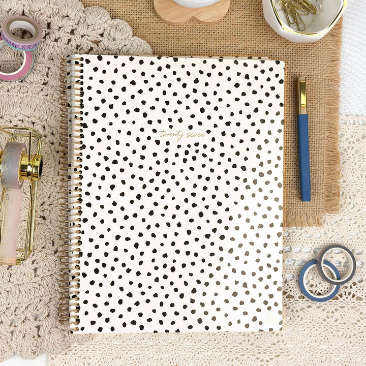 A spiral-bound planner or notebook with a white cover covered in small black polka dots, shown in a styled flat-lay with a blue pen, washi tapes, and other stationery around it.