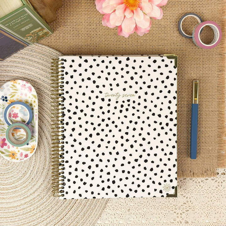 A spiral-bound planner or notebook with a white cover covered in small black polka dots and gold-toned lettering near the top center, styled in a flat lay on a burlap surface with a pink flower, washi tape, a blue pen, and other stationery props.
