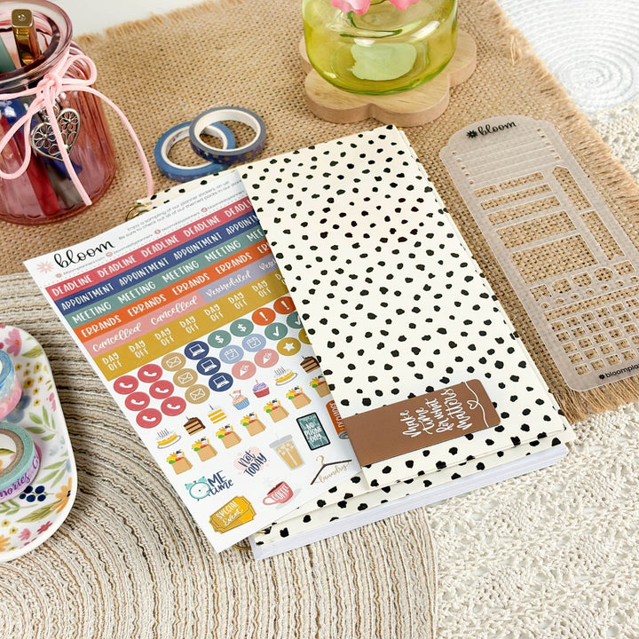 A planner sticker sheet and matching sticker pad from Bloom are displayed on a styled flat-lay surface with a white notebook featuring black polka dots, a clear plastic divider, and decorative accessories.