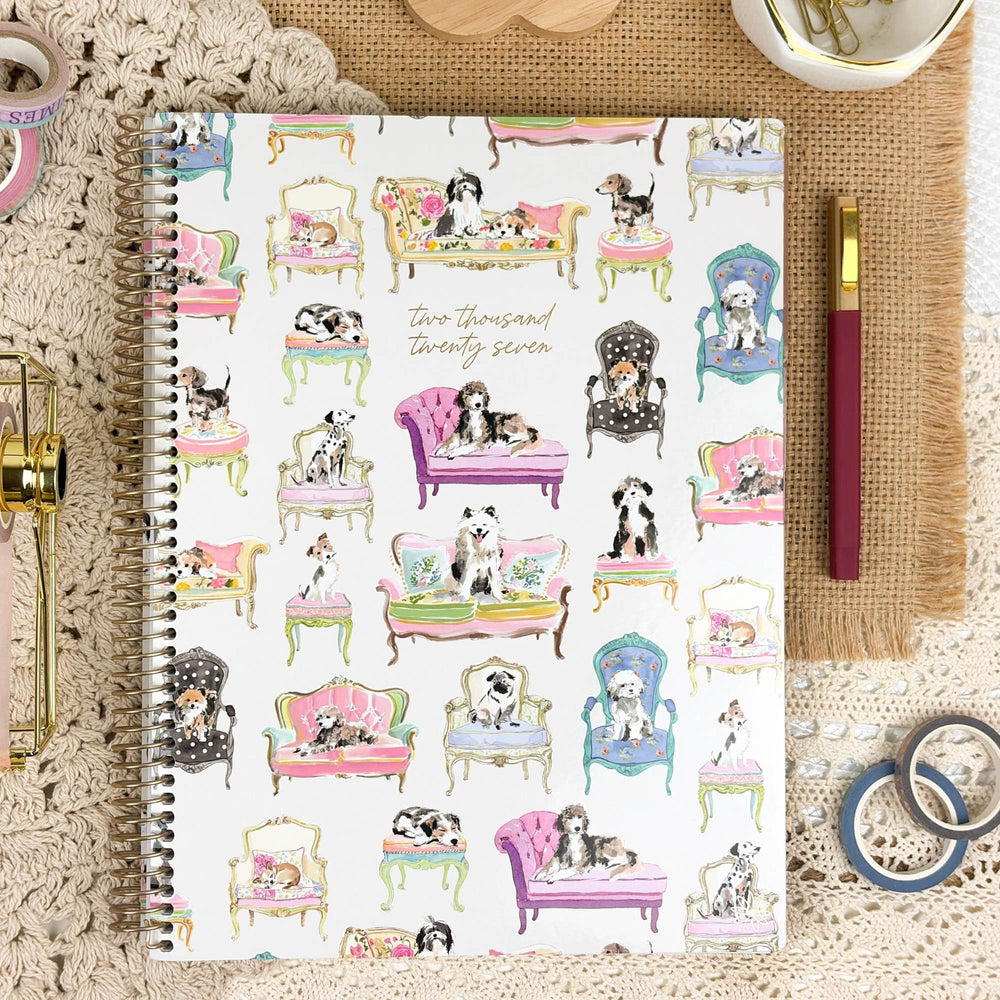 A spiral-bound notebook with a white cover decorated with colorful illustrations of dogs sitting in various vintage-style chairs and sofas, displayed in a styled flat-lay with a pen, washi tape, and a crochet blanket. - hover