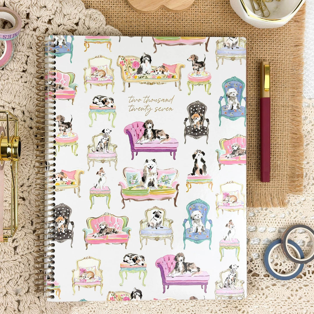 A spiral-bound notebook with a white cover decorated with colorful illustrations of dogs sitting in various vintage-style chairs and sofas, displayed in a styled flat-lay with a pen, washi tape, and a crochet blanket.
