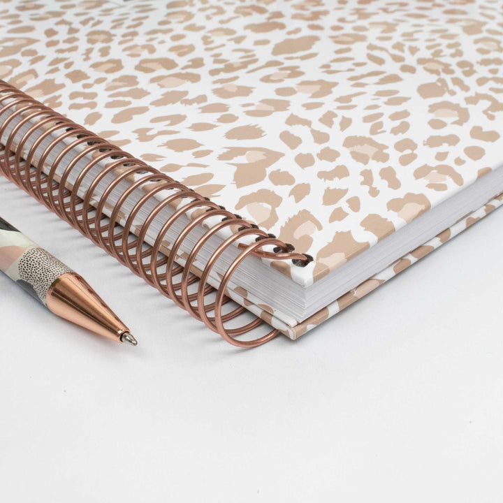 A spiral-bound notebook with a beige and white leopard print cover, shown on a white background next to a matching rose gold pen.