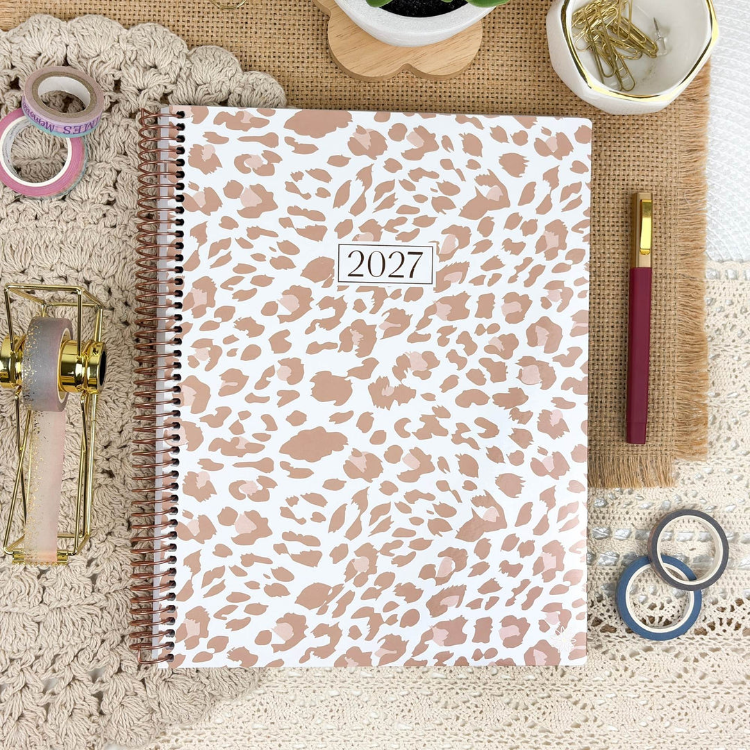 A 2027 spiral-bound planner with a white cover featuring a dusty rose leopard print pattern, styled in a flat-lay arrangement with stationery accessories on a burlap and lace background.