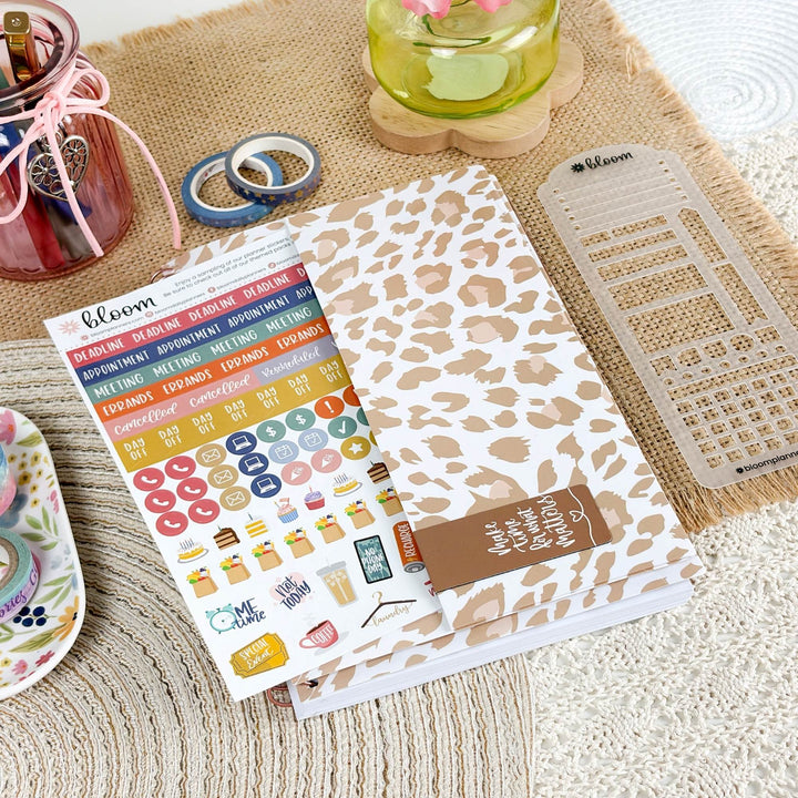 A planner set styled on a woven beige mat, featuring a leopard-print planner cover, a sheet of colorful planner stickers, a clear plastic calendar grid, and a small jar of pink ribbon.