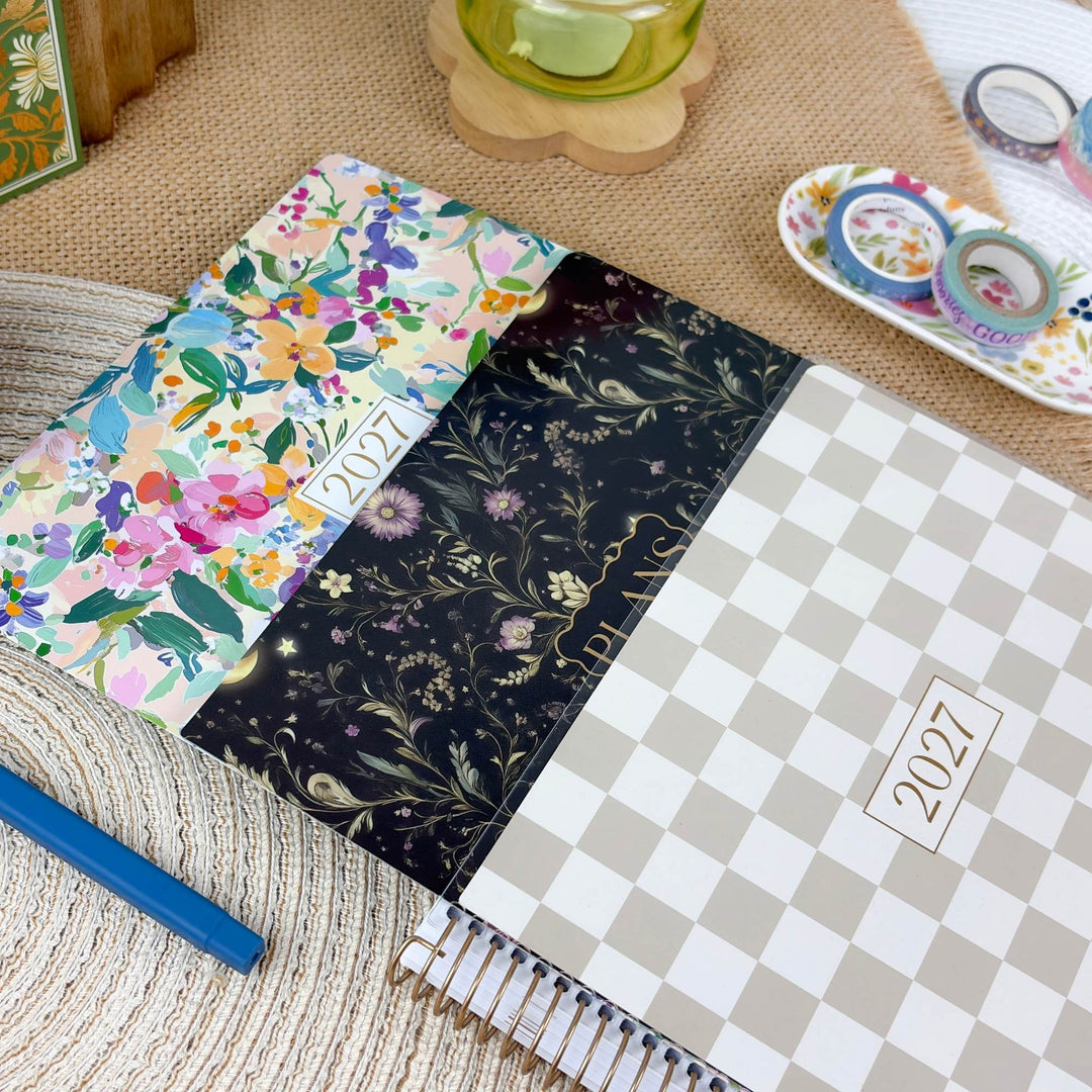 A spiral-bound 2027 planner shown open on a woven mat, with three floral-patterned covers visible: a multicolor floral cover, a black floral cover with gold details, and a beige-and-white checkerboard cover. A blue pen, washi tape rolls, and other desk props are arranged around it.