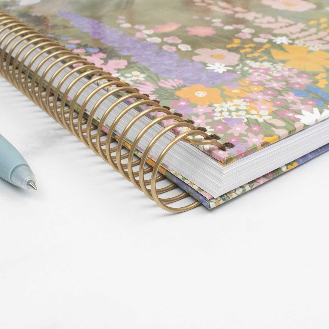 A close-up of a spiral-bound notebook with a multicolor floral cover and a gold-tone spiral binding, shown on a white surface with a light blue pen partially visible at the left edge.