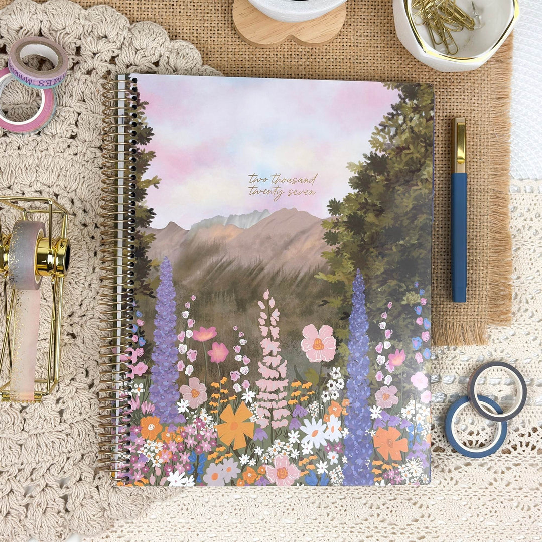 A spiral-bound notebook with a colorful watercolor floral and mountain cover design, styled in a flat lay on a lace cloth with washi tape, a gold pen, and other stationery props.
