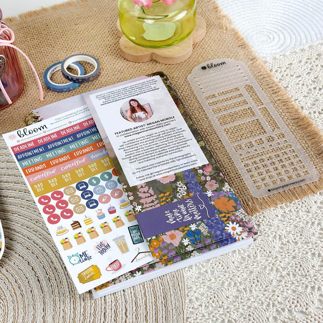 A styled flat-lay of a Bloom planner sticker sheet set on a woven beige mat, featuring colorful category stickers, a floral-themed insert sheet, and a clear plastic grid template.
