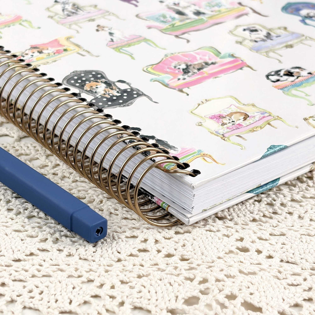 A spiral-bound notebook with a white cover decorated with colorful illustrations of dogs in vintage furniture, shown on a cream lace doily next to a blue pen.