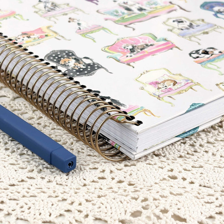A spiral-bound notebook with a white cover decorated with colorful illustrations of dogs in vintage furniture, shown on a cream lace doily next to a blue pen.