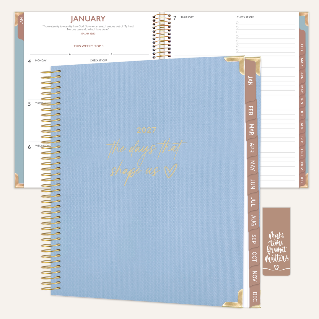 A spiral-bound planner shown open and closed on a light cream background. The closed cover is light blue with gold foil script reading '2027 the days that sleep us' and gold corner accents.