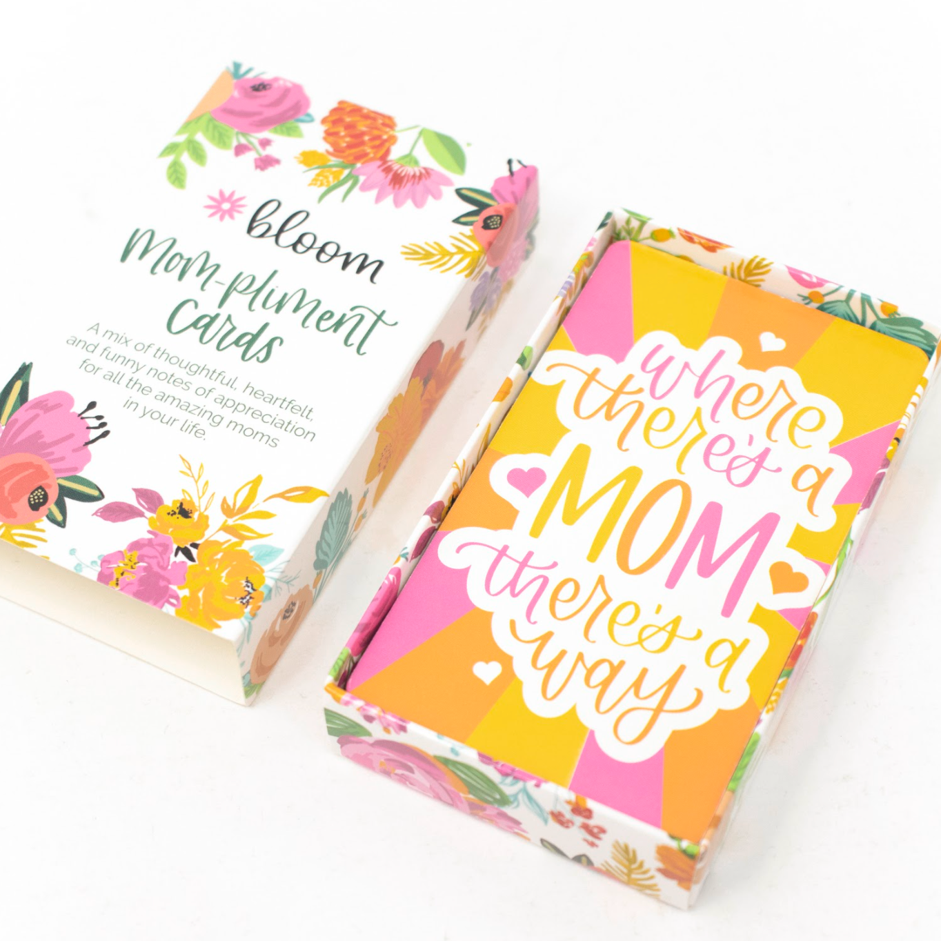 A colorful box labeled bloom Mompliment Cards is open, revealing cards inside. The visible card reads, Where theres a mom theres a way. The box and cards feature bright floral designs.