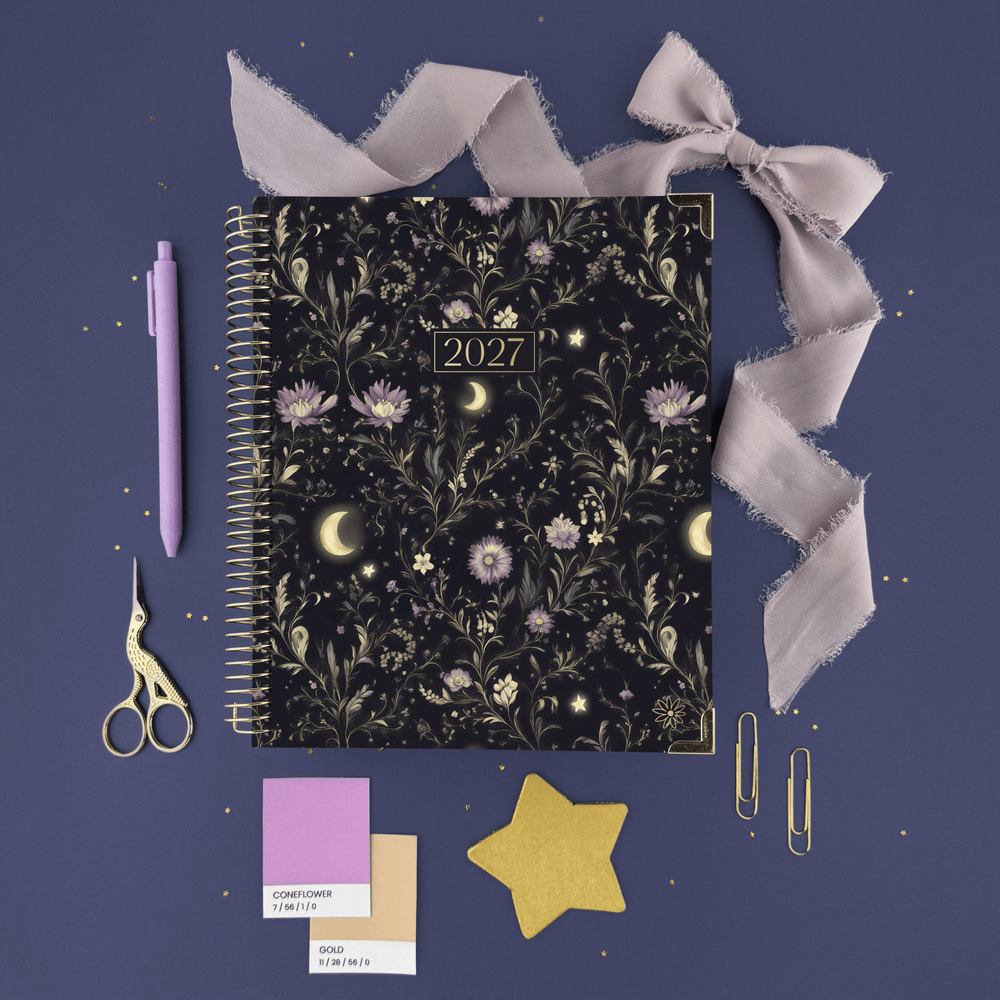 A 2027 spiral-bound planner with a dark floral and celestial pattern is displayed in a styled flat-lay on a dusty purple background. The planner is surrounded by coordinating lavender stationery items, including a pen, ribbon, scissors, paper clips, and color swatches. - hover