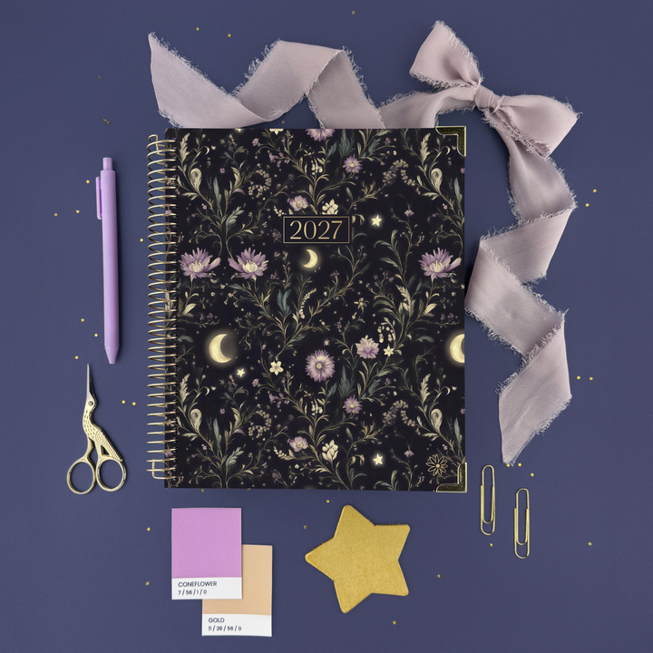 A 2027 spiral-bound planner with a dark floral and celestial pattern is displayed in a styled flat-lay on a dusty purple background. The planner is surrounded by coordinating lavender stationery items, including a pen, ribbon, scissors, paper clips, and color swatches.