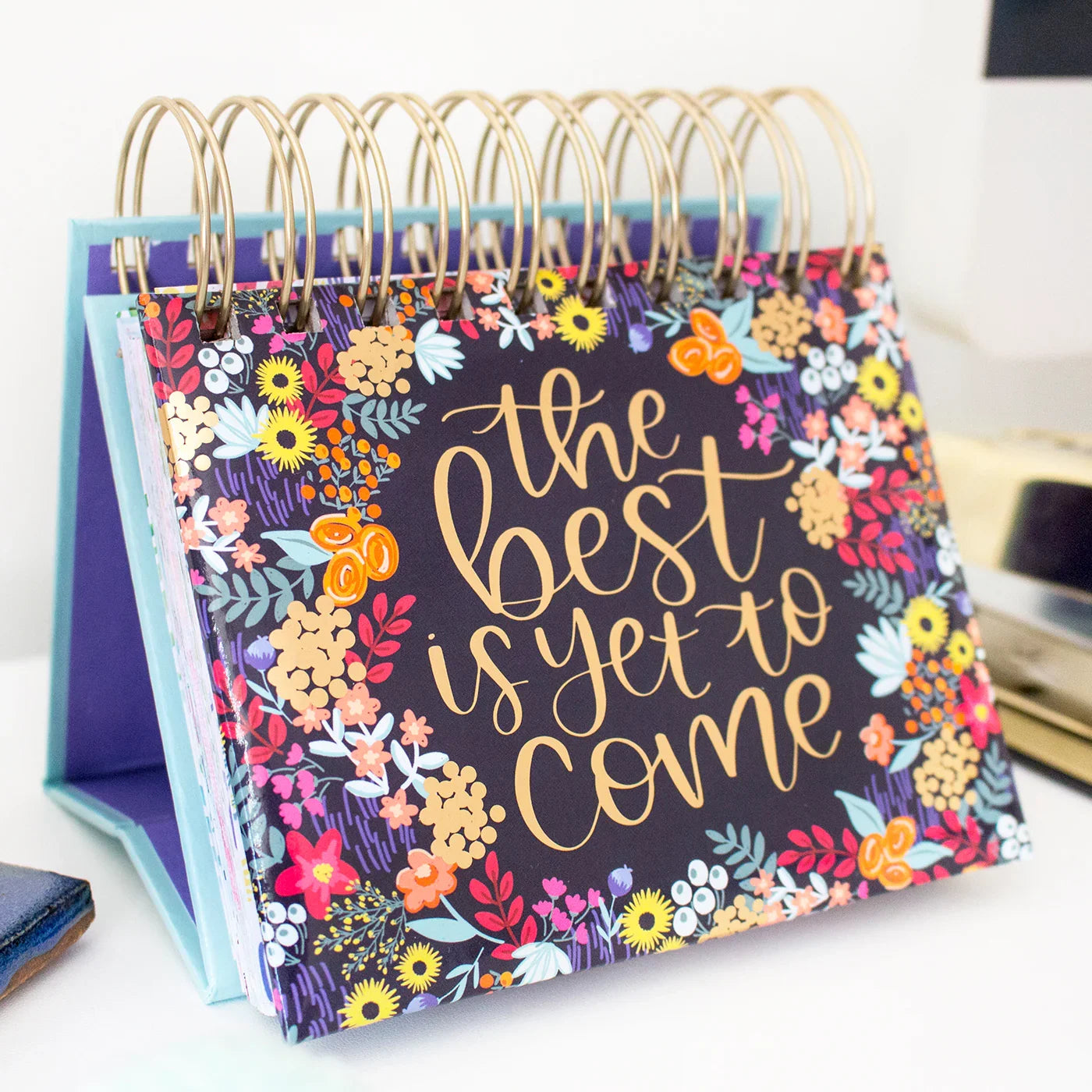 The bloomdailyplanners Inspirational Perpetual Desk Easel features a vibrant floral border, a spiral binding, and "The Best is Yet to Come" in large yellow script on the cover.